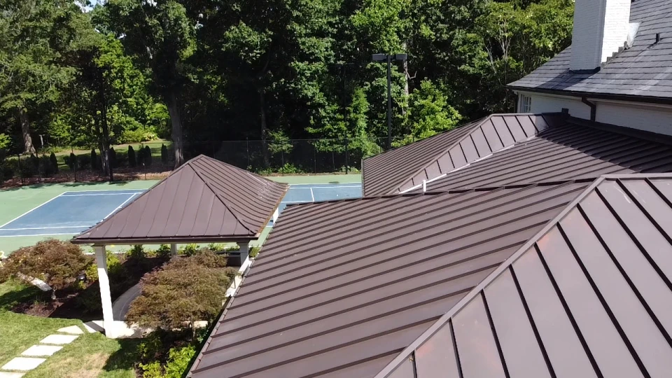 Dark brown metal standing seam roofing on a residential building next to a outdoor tennis court surrounded by trees.